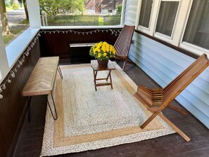 Un porche abrité avec un banc en bois, une petite table et une chaise. Un pot de fleurs contenant des fleurs jaunes est posé sur la table.