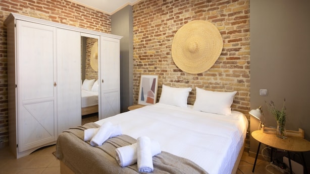 A hotel room with a brick wall, a large bed with white linens, a wardrobe, and a round hat on the wall. - Villa Rosa Suite by CorfuEscapes (Corfu)