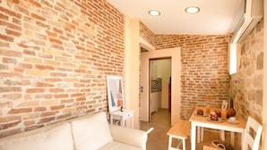 A room with a brick wall, a white sofa, a small table with a vase, and a door leading to a kitchen. - Villa Rosa Suite by CorfuEscapes (Corfu)