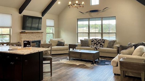 A spacious living room with a high ceiling, a fireplace, a large window, and a dining area with a kitchen island. - Contemporary Waterfront Ranch Villa at Twin Canyons Longhorn Ranch (Granbury)
