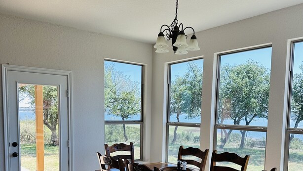 Un comedor con mesa y sillas de madera, ventanas amplias con vistas a árboles y una masa de agua, y una puerta que da al exterior.
