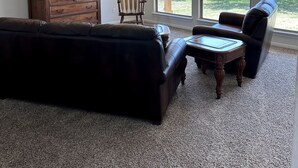Sala de estar con un sofá de cuero oscuro, una silla de madera, una mesita de madera pequeña y un mueble de madera grande. Hay ventanas amplias con vistas a árboles y un edificio.