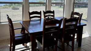 Un comedor con una mesa grande y sillas, ventanas amplias con vistas a un lago y árboles, y una lámpara de araña.