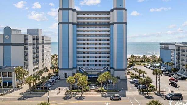 A hotel with a central entrance, multiple floors, and a parking lot with cars and palm trees. - Enjoy the Breathtaking View from Your Private Oceanfront Balcony (Myrtle Beach)