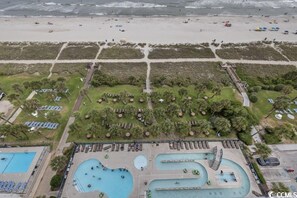 Vista aérea de um resort à beira-mar, com várias piscinas, espreguiçadeiras e uma área de praia com guarda-sóis e pessoas.