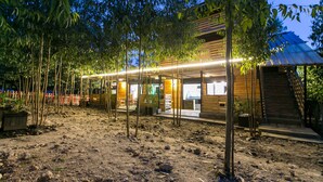 Un edificio rustico in legno con una zona esterna coperta, circondato da alberi e terreno sterrato.