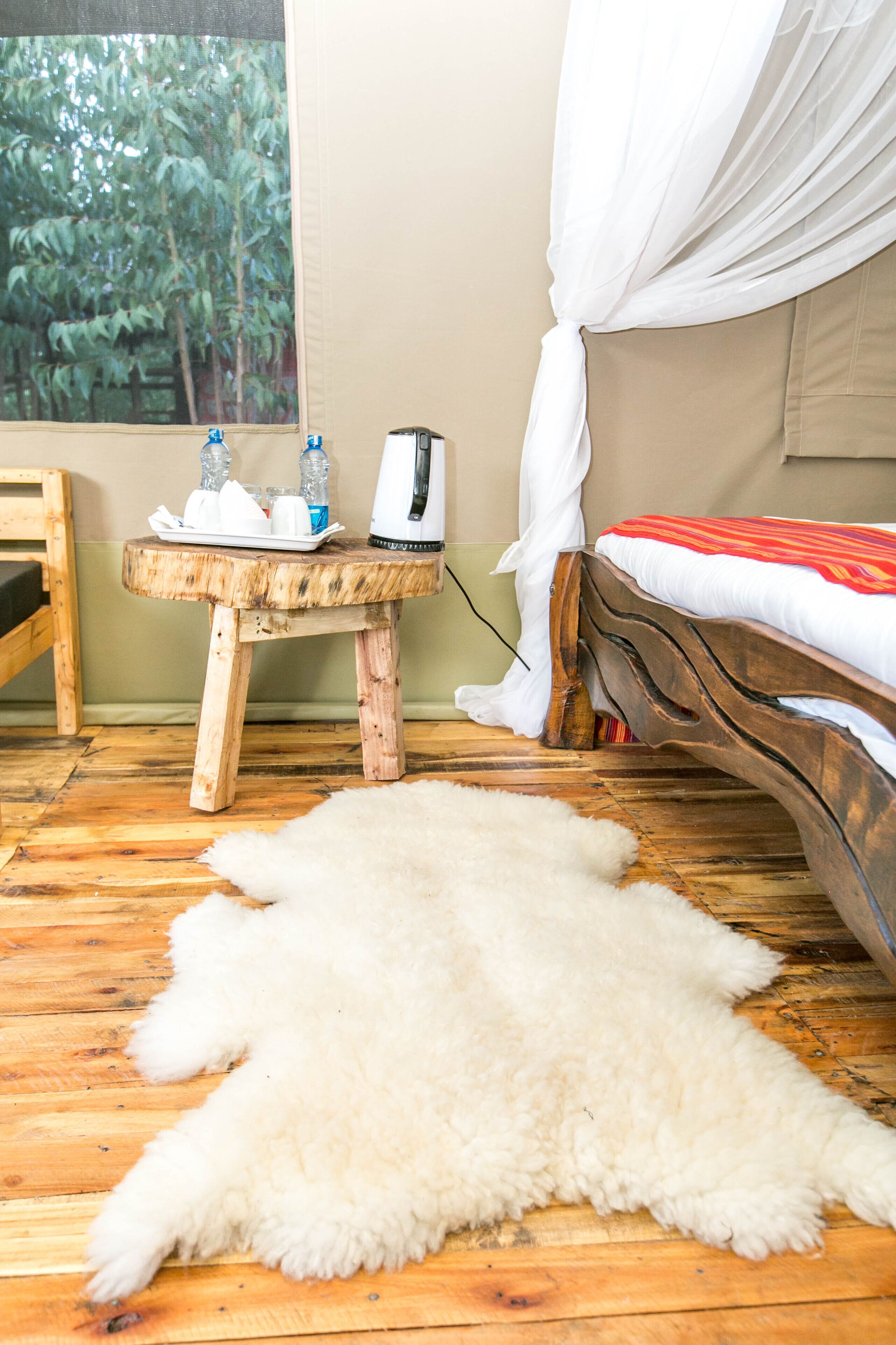 A room with a wooden floor, a white fur rug, a bed with a white canopy, a small wooden table with a kettle and bottles, and a view of trees through a window.