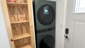 Une buanderie équipée d’un lave-linge et d’un sèche-linge superposés, d’une étagère en bois sur laquelle se trouve une boîte de lessive Tide, et d’une porte blanche avec une fenêtre.
