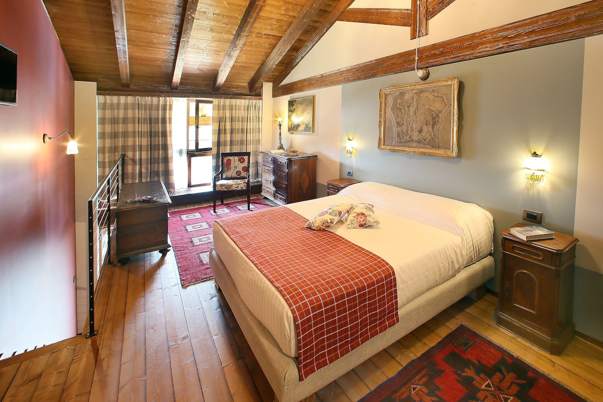 Un dormitorio con techo de madera, una cama con manta roja, una mesita de noche, un aparador y una ventana con cortinas.