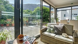 A modern room with large windows, a wicker sofa with cushions, a small table with a newspaper and glasses, and potted plants. - Solarium Villa by Atlantic Holiday (Machico)