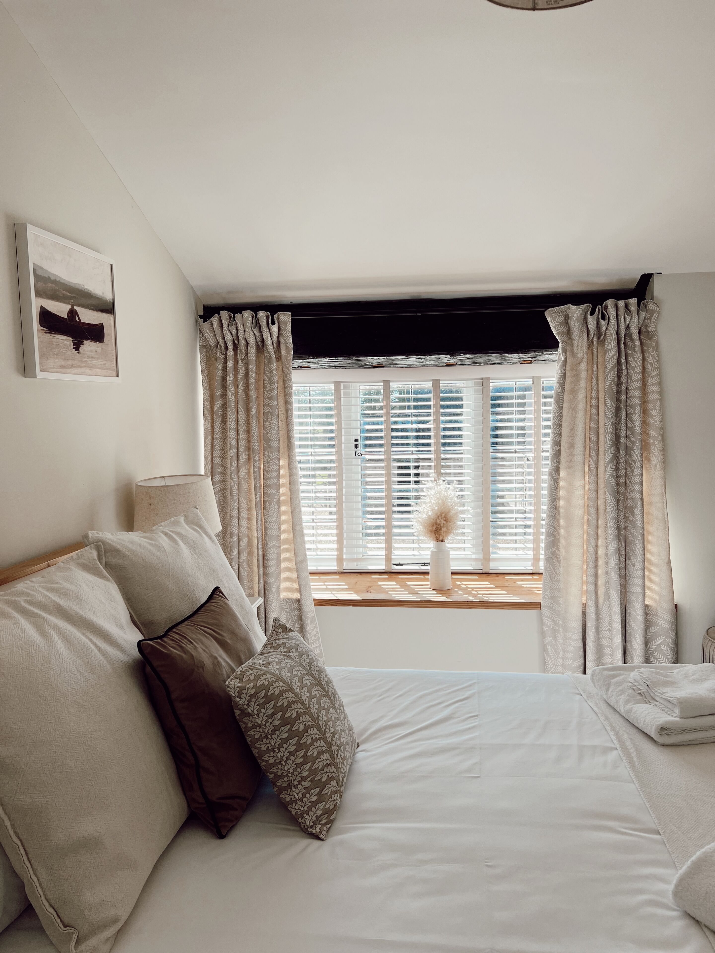 A bedroom with a bed, pillows, a window with curtains, a bedside table, and a wall-mounted lamp.