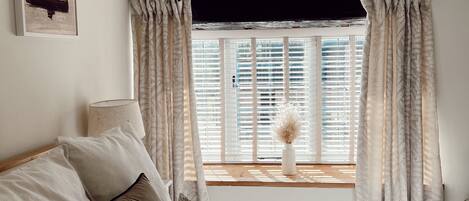 A bedroom with a bed, pillows, a window with curtains, a bedside table, and a wall-mounted lamp.