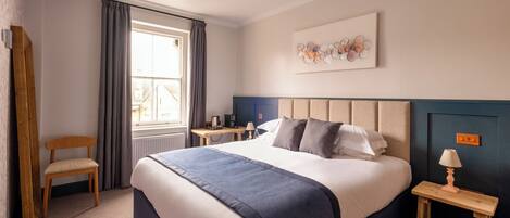 A hotel room with a large bed, a wooden chair, a small table with a lamp, and a window with curtains.