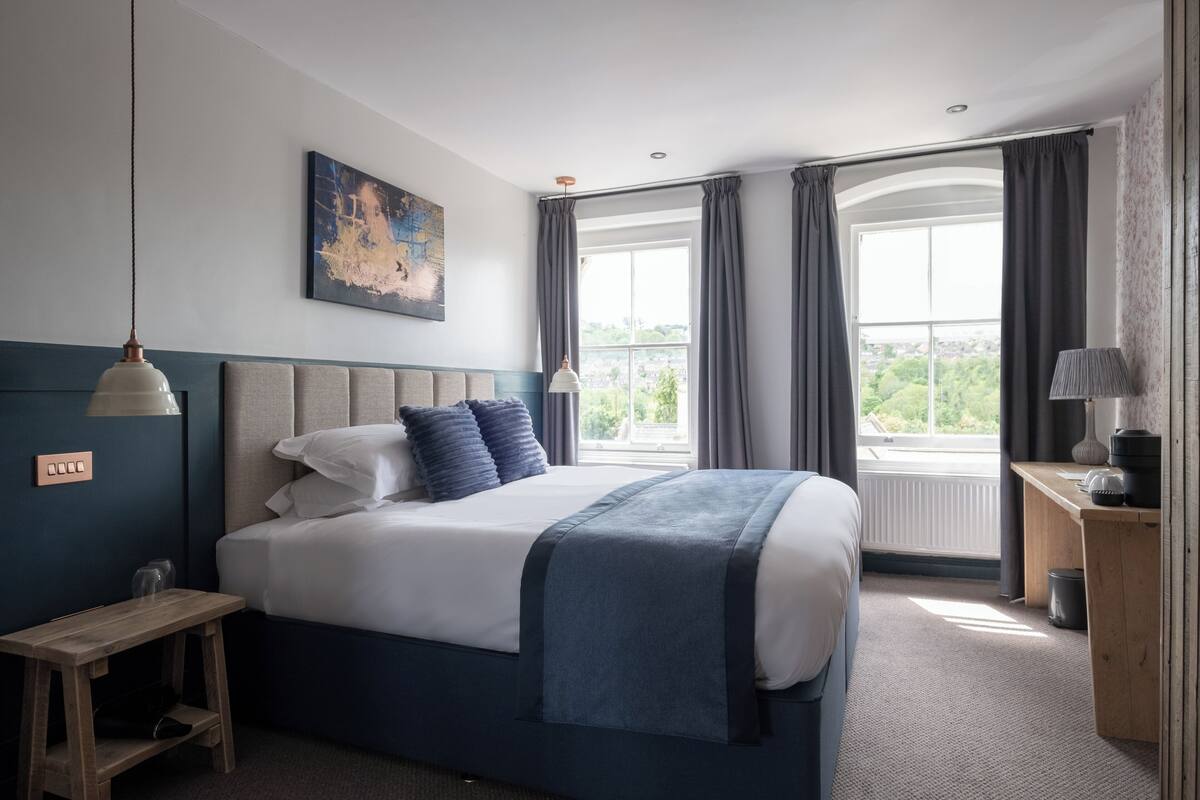 A bedroom with a large bed, a bedside table, a lamp, and a view of the outdoors through a window.