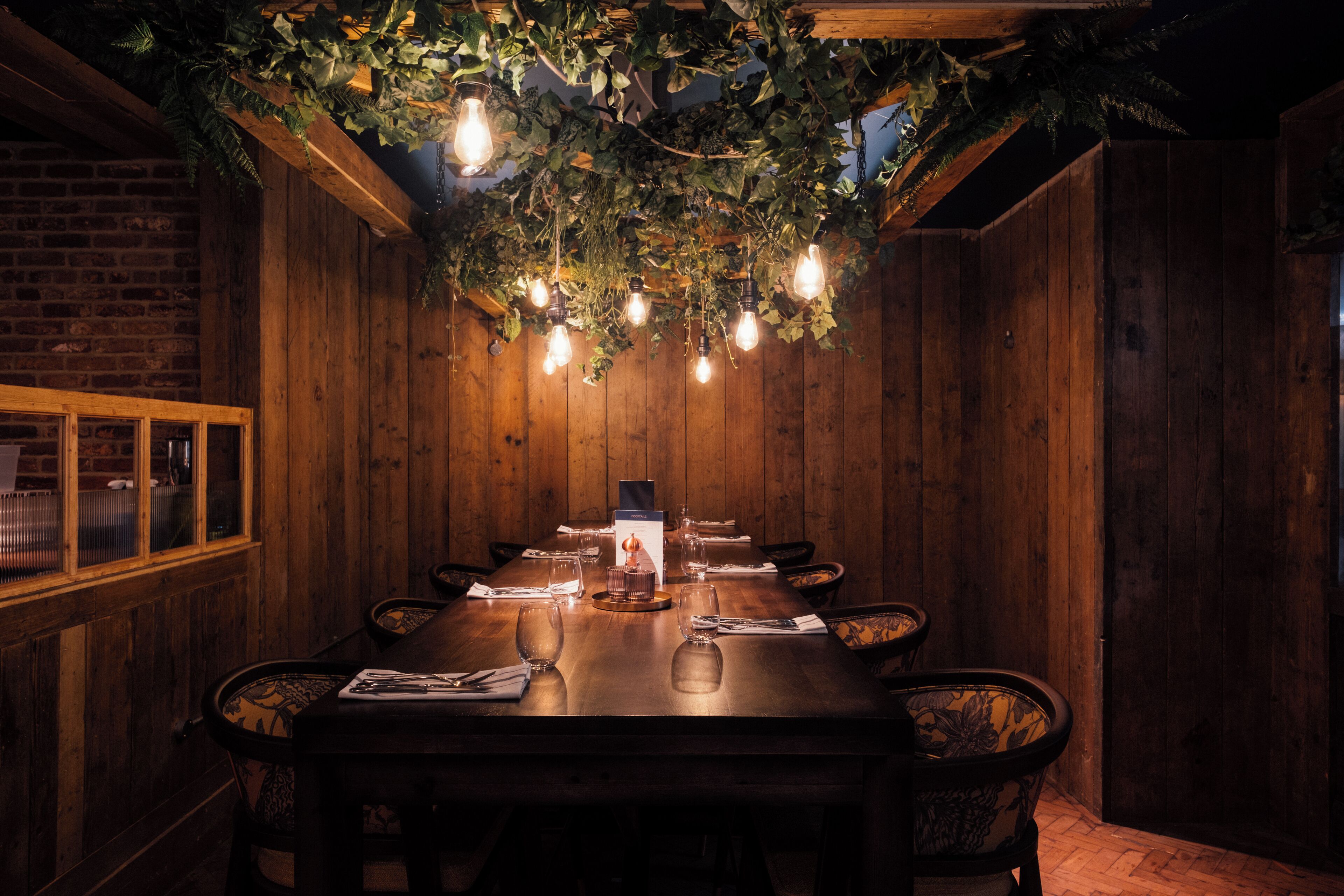A wooden dining table with chairs, lit by hanging bulbs, surrounded by wooden walls and a brick wall.