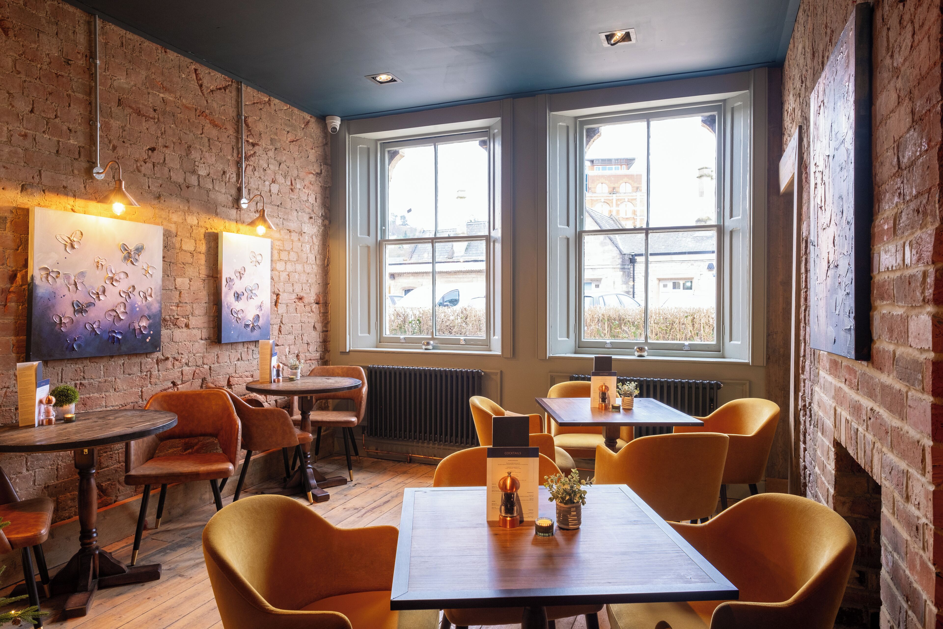 A cozy restaurant with brick walls, large windows, and orange chairs. Tables are set with menus and small plants.