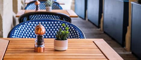 A covered outdoor seating area with wooden tables and blue patterned chairs, featuring a small potted plant and a copper salt shaker.