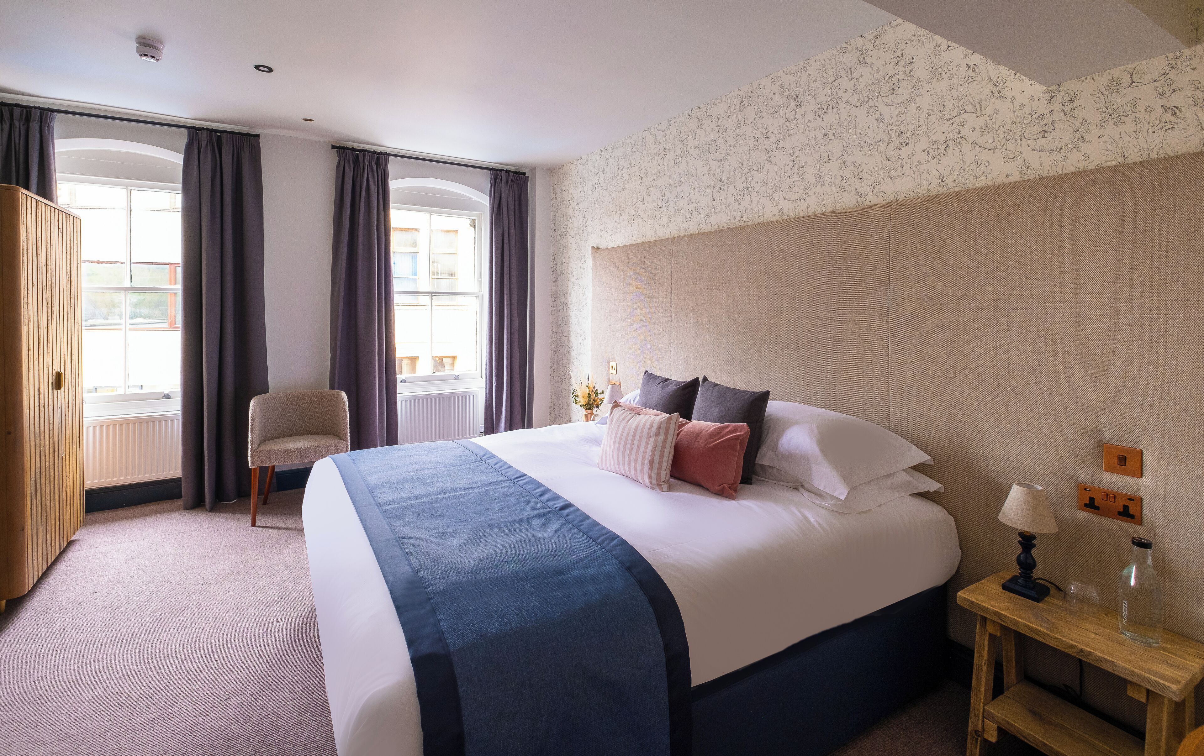 A hotel room with a large bed, a chair, a bedside table with a lamp, and a view of the outside through a window.
