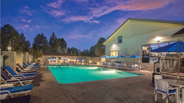 A swimming pool with lounge chairs and umbrellas, surrounded by a building with a covered area and a view of trees. - 1Bed/1Bath - Gorgeous Flagstaff Location with Pools, Golf, and More! (Flagstaff)