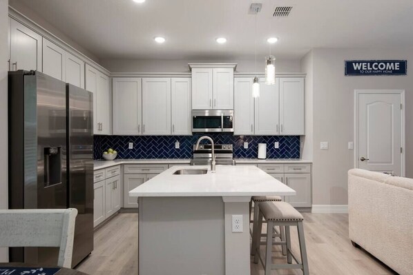 Une cuisine moderne avec des meubles blancs, un îlot central, des appareils en acier inoxydable et un pare-feu bleu.