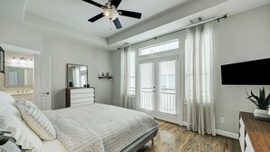 A bedroom with a bed, ceiling fan, large windows with blinds, a dresser, and a TV on the wall.