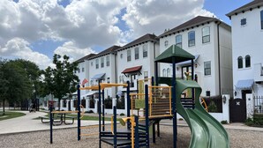 A playground with a slide, climbing structures, and a picnic table in front of a row of white houses with balconies.