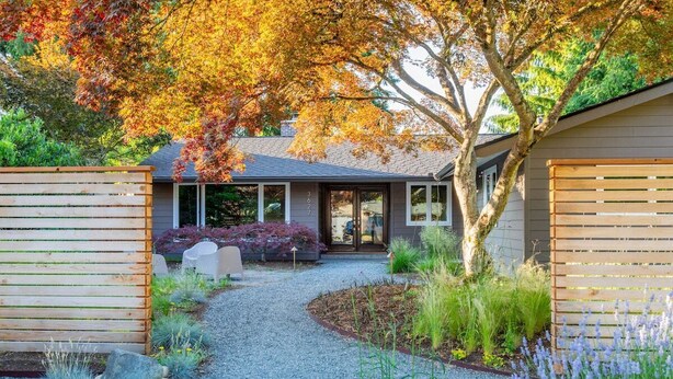Una casa moderna con camino de grava, cerca de madera y jardín con diversas plantas y un árbol de hojas otoñales.