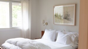 Una cama con ropa blanca impecable en una habitación con un ventanal grande, una lámpara de pared y un cuadro enmarcado.