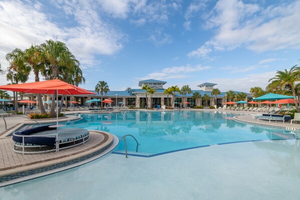A large outdoor swimming pool with lounge chairs and umbrellas, surrounded by palm trees and a building with a blue roof.