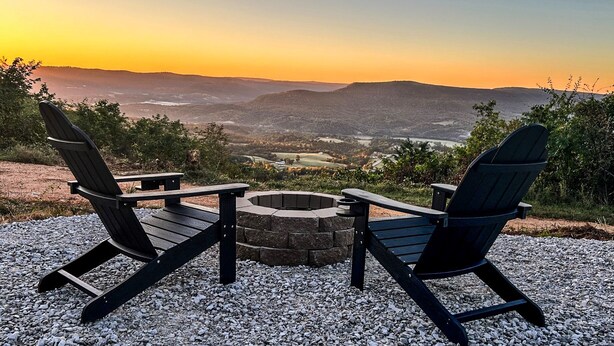 Dos sillas Adirondack mirando un hogar a leña de piedra con una vista panorámica de un valle y montañas al atardecer.