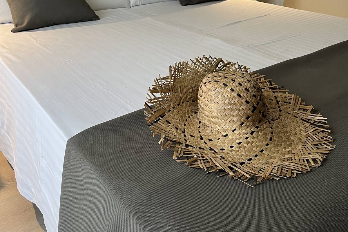 A neatly made bed with white linens and a dark gray bedspread, a wooden headboard, and a bedside table with a lamp and a small potted plant.