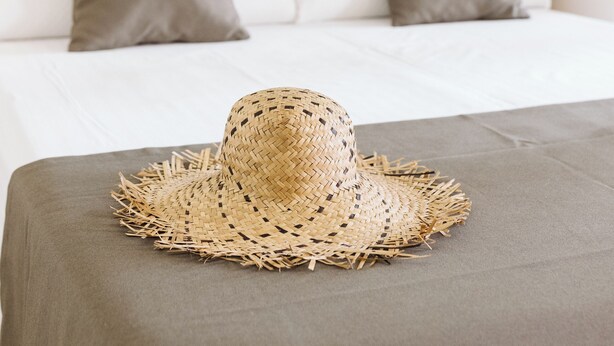 Una cama con un sombrero tejido en el borde, cabecero de madera y almohadas grises.