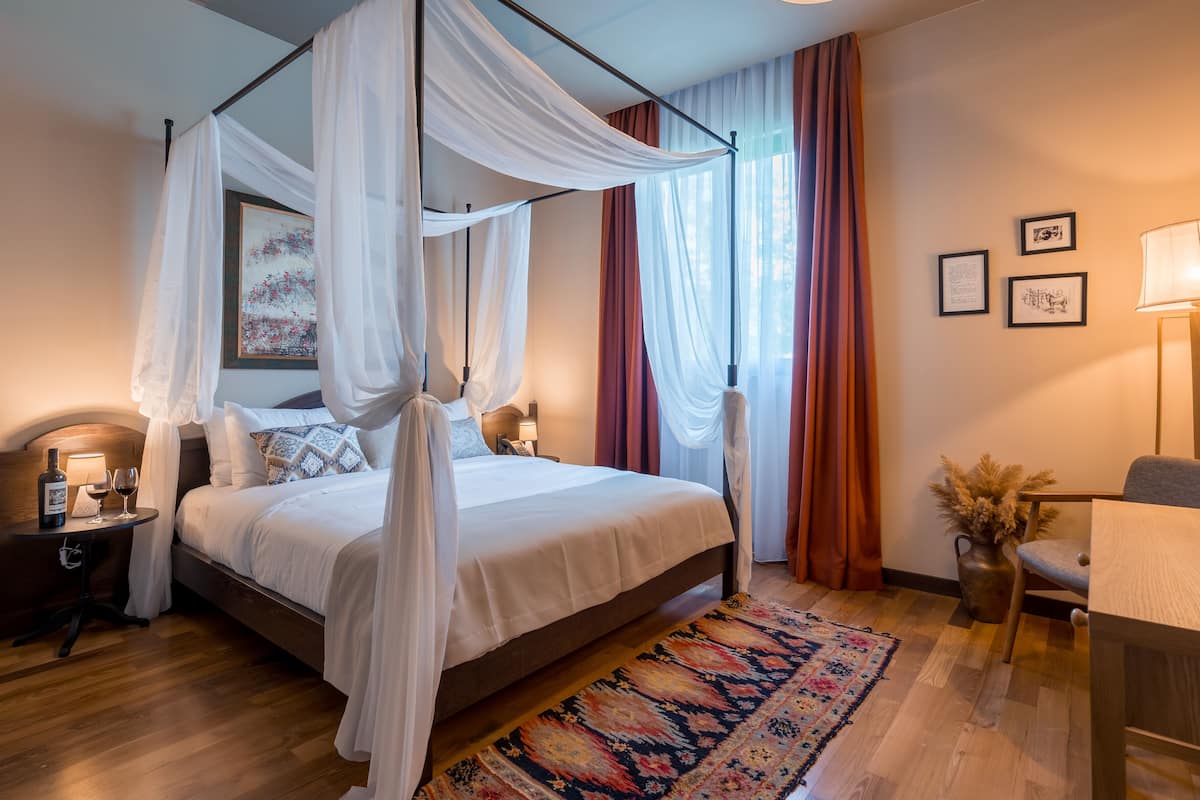 A bedroom with a four-poster bed, a bedside table with a lamp, a chair, a desk, and a large window with curtains.