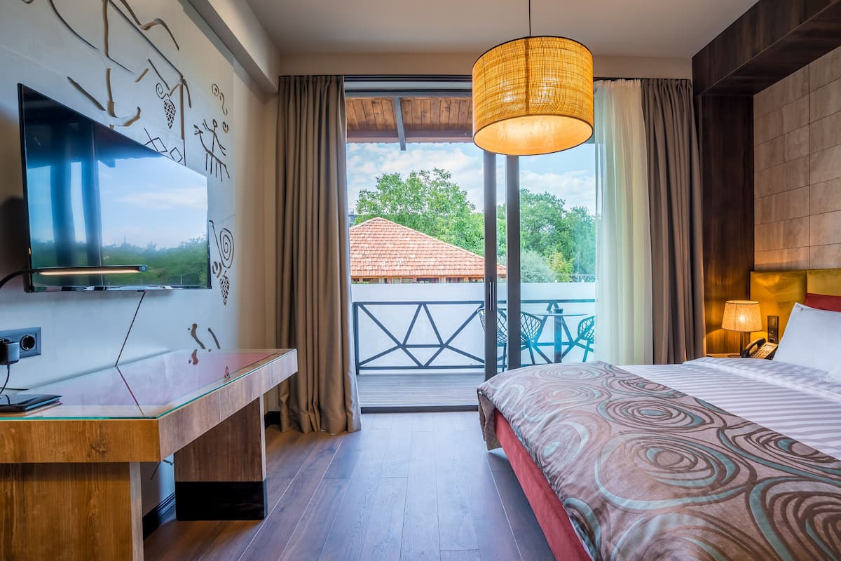 A hotel room with a bed, a desk, a TV, and a balcony with a view of trees and a building.