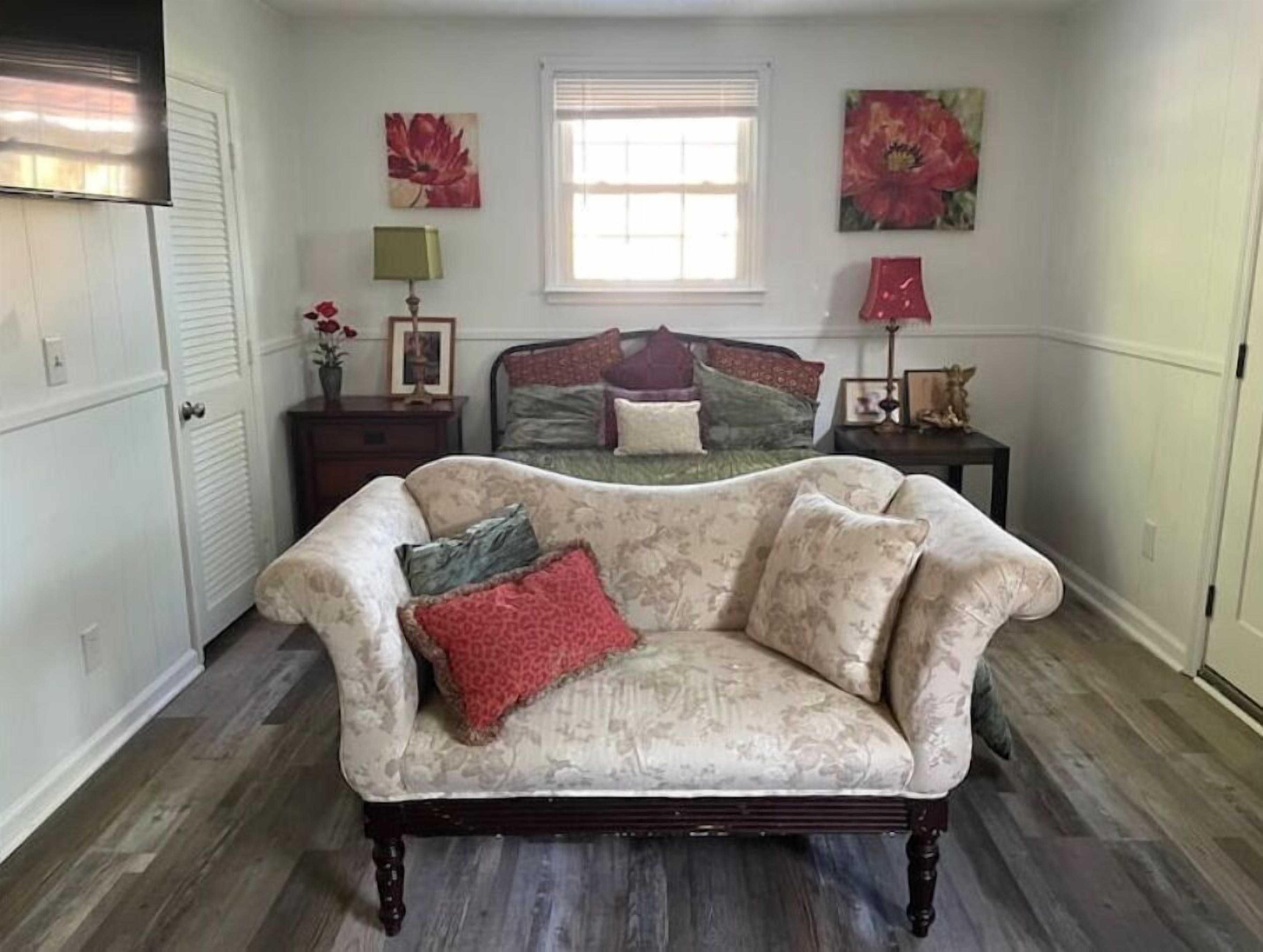 A bedroom with a floral-patterned sofa, a bed with multiple pillows, a window with blinds, and two wall-mounted lamps.