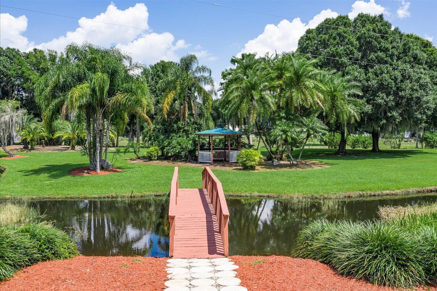 A wooden bridge over a pond with a gazebo in the background surrounded by lush greenery.