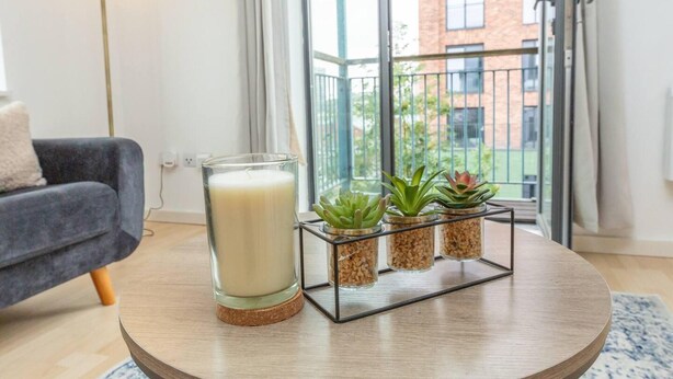 Une table ronde en bois, sur laquelle se trouvent une bougie dans un verre, un verre de lait et trois plantes grasses en pot.