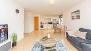 Un salon moderne avec un canapé, une table basse et un téléviseur. Au fond, on aperçoit une cuisine avec des meubles blancs et un coin repas comprenant une table et des chaises.