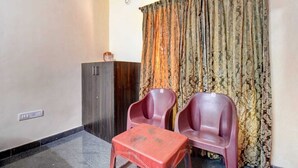 Una habitación con un armario de madera oscura, dos sillas de plástico, una mesita roja y una cortina estampada.