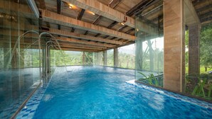 Une piscine intérieure avec des parois en verre transparent, des poutres en bois au plafond et une vue sur la verdure environnante.