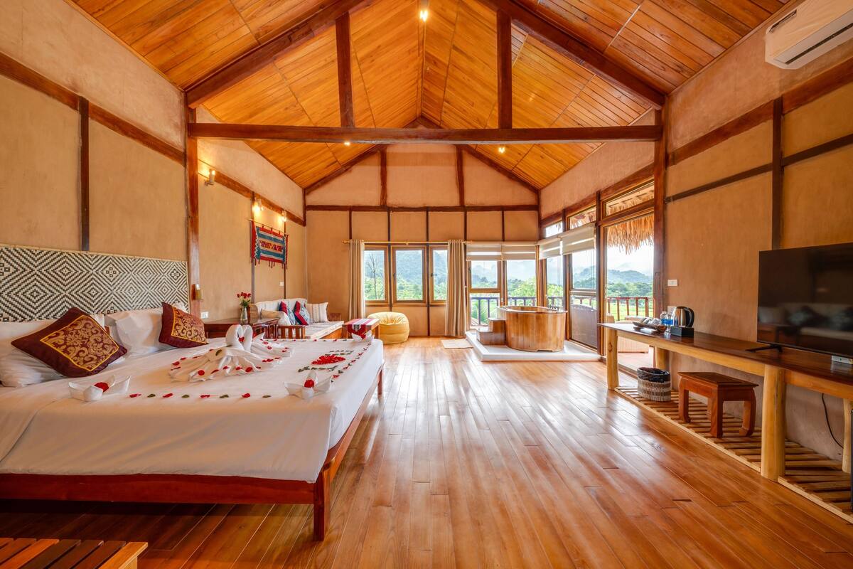 Ein geräumiges Zimmer mit einem großen Bett, Holzboden, einem Fernseher und Blick ins Freie.