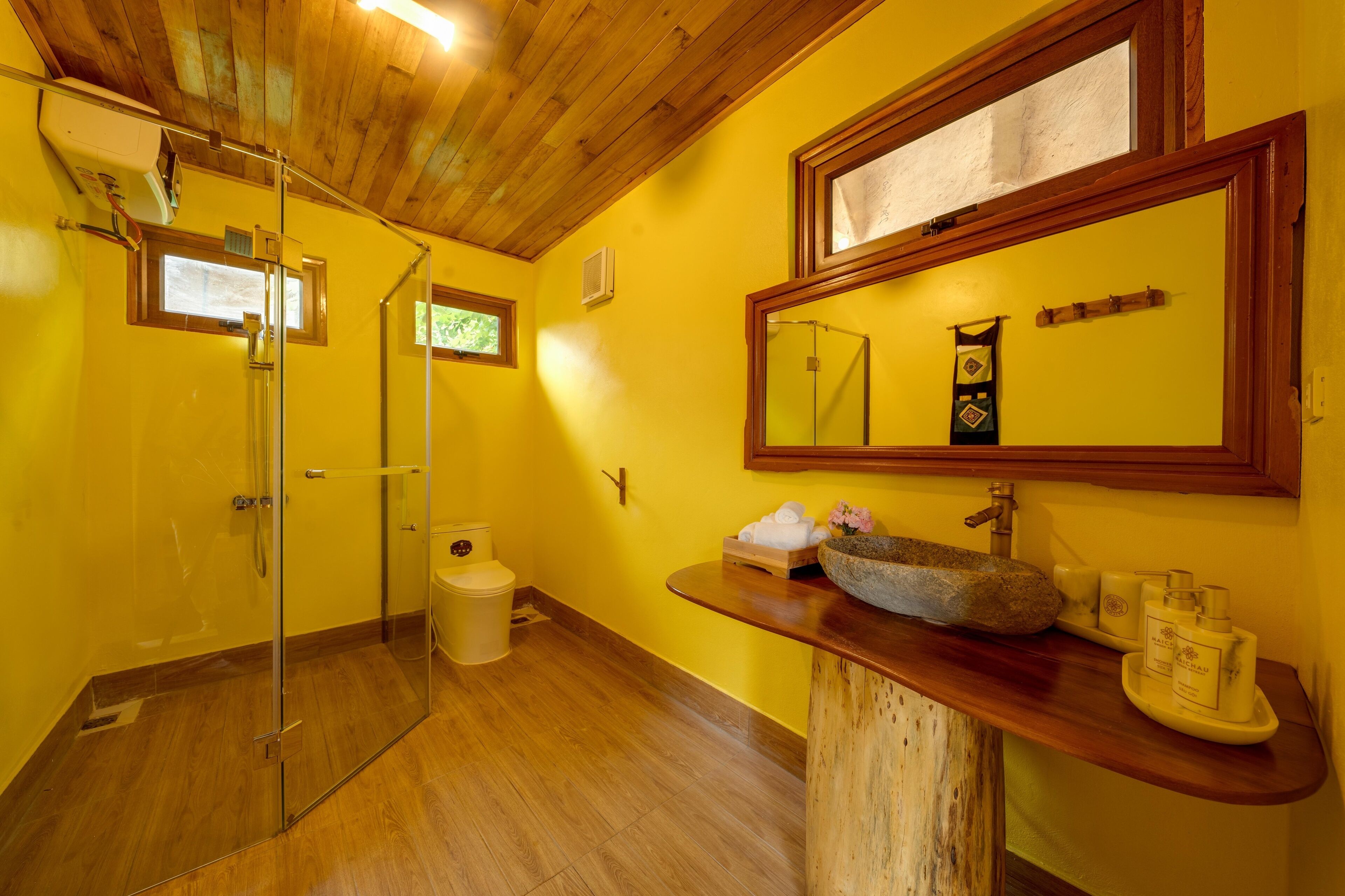 Un baño con techo de madera, paredes amarillas, cabina de ducha de vidrio, tocador de madera y espejo sobre el lavamanos.