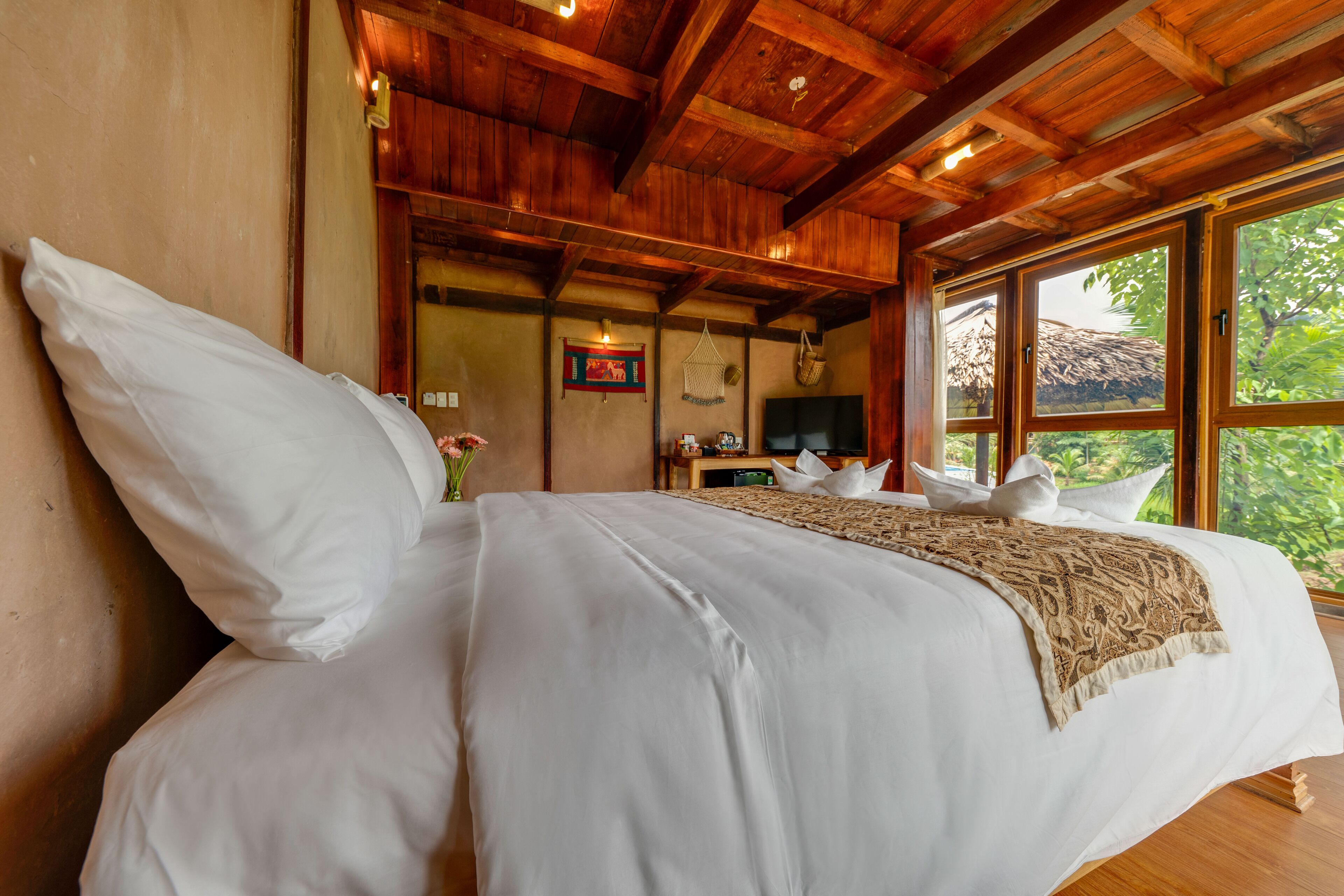 A large bed with white linens and a beige throw blanket, wooden ceiling beams, a small table with a television, and a window with a view of greenery.