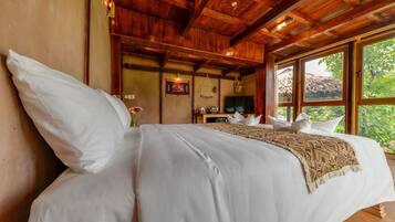 A large bed with white linens and a beige throw blanket, wooden ceiling beams, a small table with a television, and a window with a view of greenery.