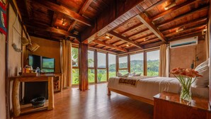 Une chambre spacieuse avec un grand lit, des poutres apparentes en bois au plafond, une télévision et une vue sur l’extérieur.