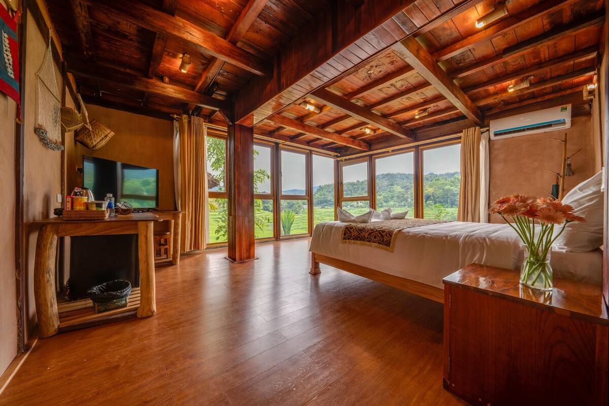 Ein geräumiges Schlafzimmer mit einem großen Bett, Holzbalkendecke, Fernseher und Blick ins Freie.