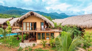 Un bungalow au toit de chaume, doté d’un balcon, d’une piscine et entouré d’une végétation luxuriante.