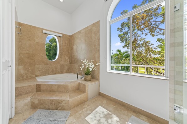 A modern bathroom with a large bathtub, a window with a view of greenery, and a small window with a view of trees.