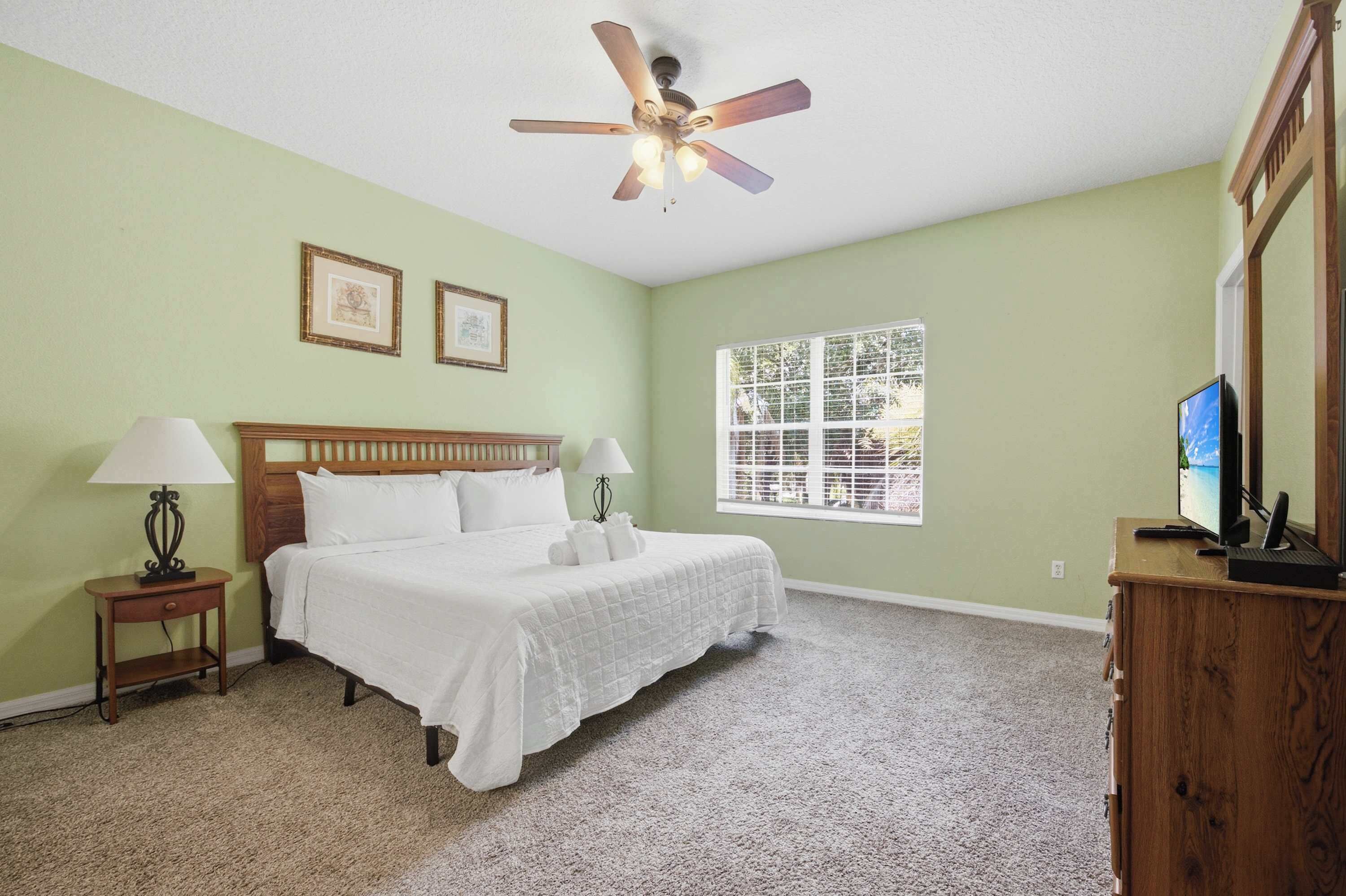 Une chambre à coucher comprenant un lit, deux tables de chevet, un ventilateur de plafond, une fenêtre et un téléviseur posé sur une commode en bois.