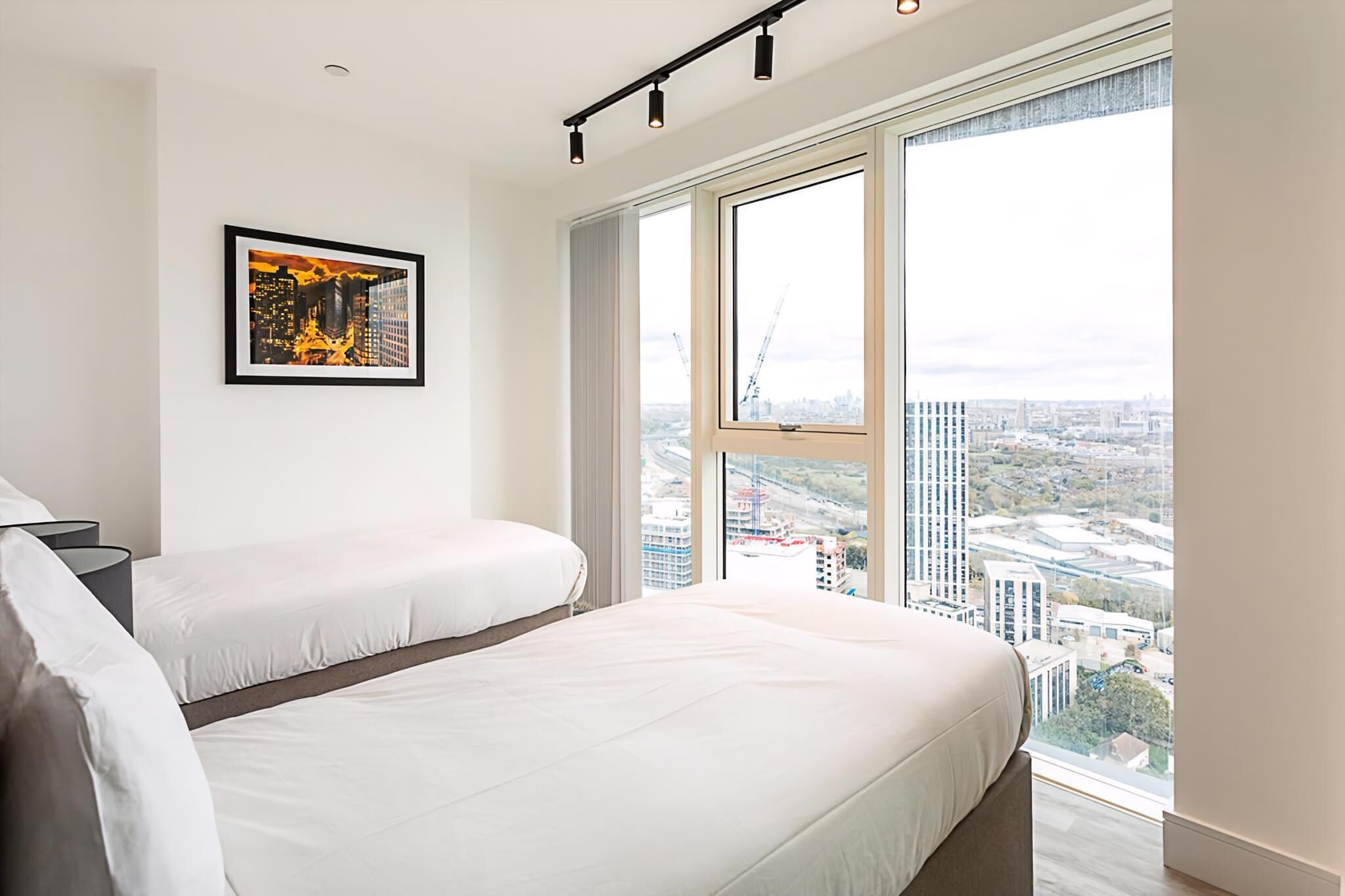 A modern hotel room with a large bed, a framed cityscape artwork, and a view of the cityscape through large windows.